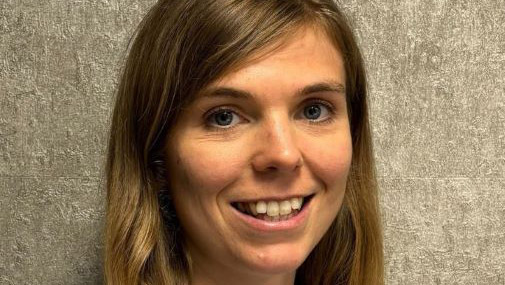 Person with long, light brown hair wearing a light blue button‑down shirt, standing against a textured gray wall.